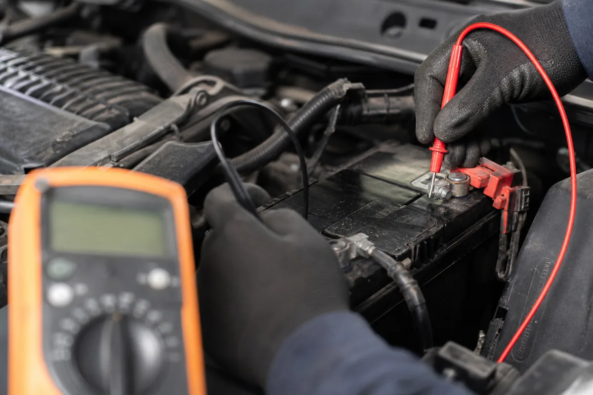 Mains gantées d&rsquo;un mécanicien testant la tension d&rsquo;une batterie de voiture au multimètre dans un atelier